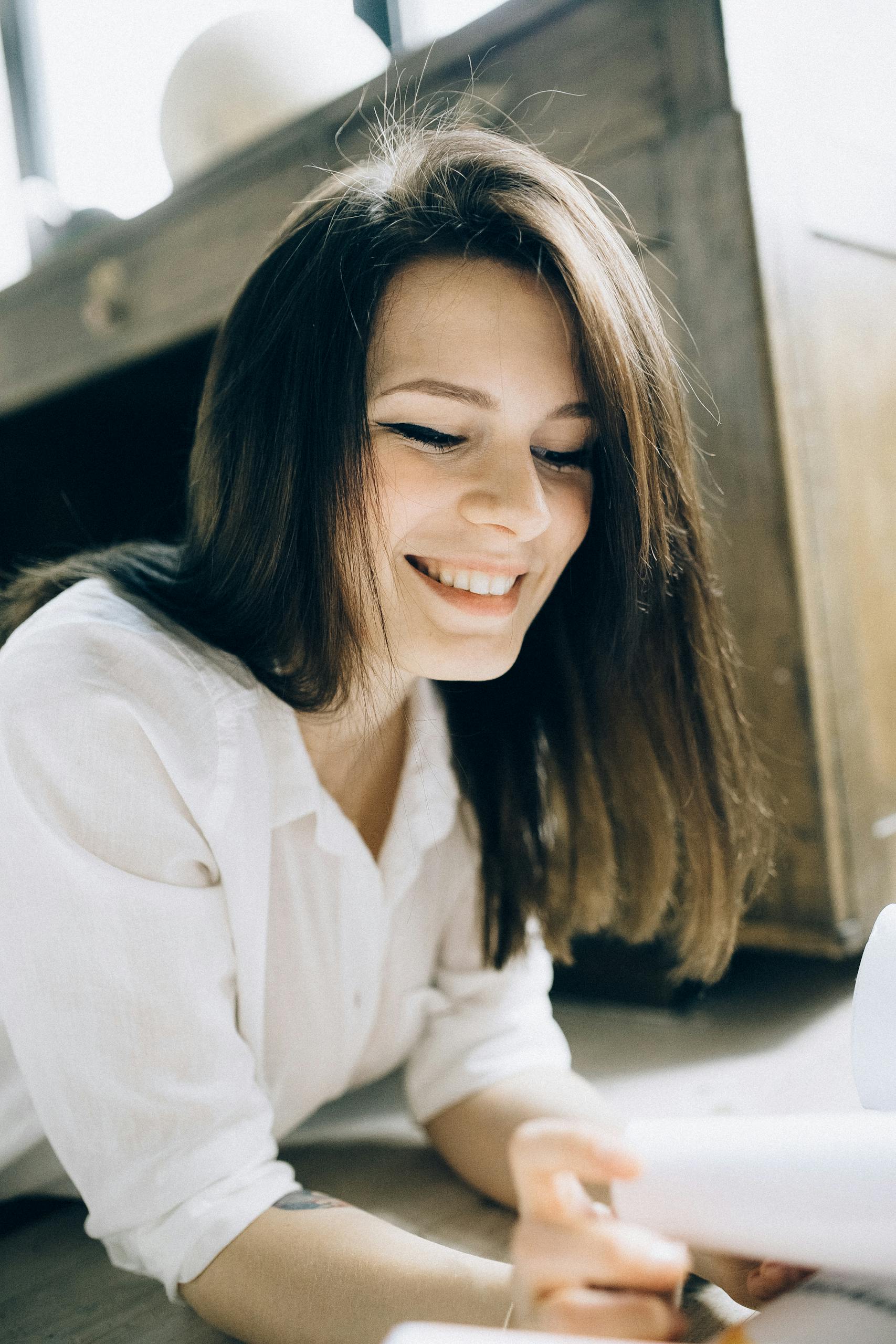 A woman smiling brightly while reading indoors on a sunny day.