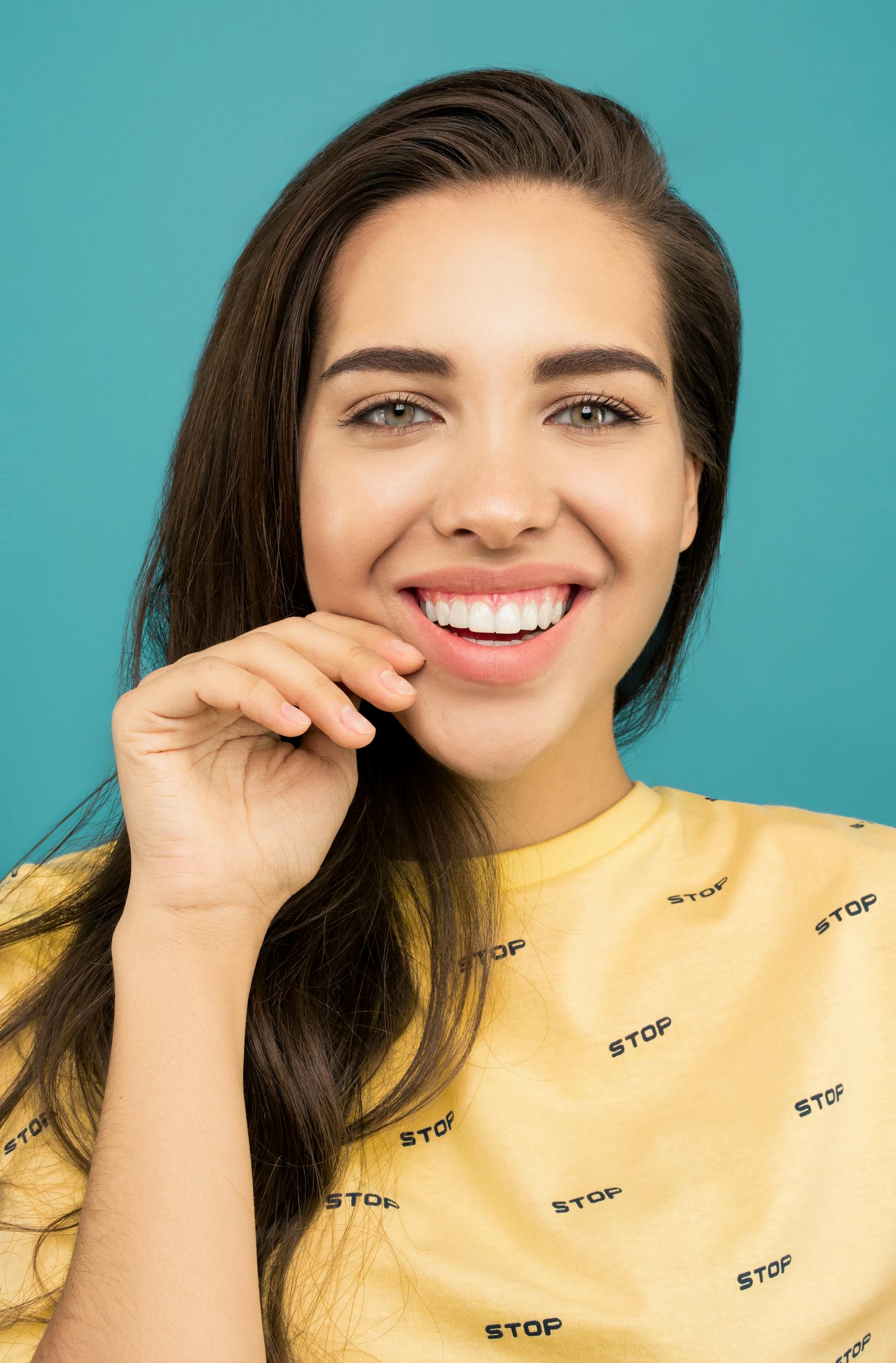 Portrait of a smiling woman with long hair wearing a yellow shirt against a turquoise background.