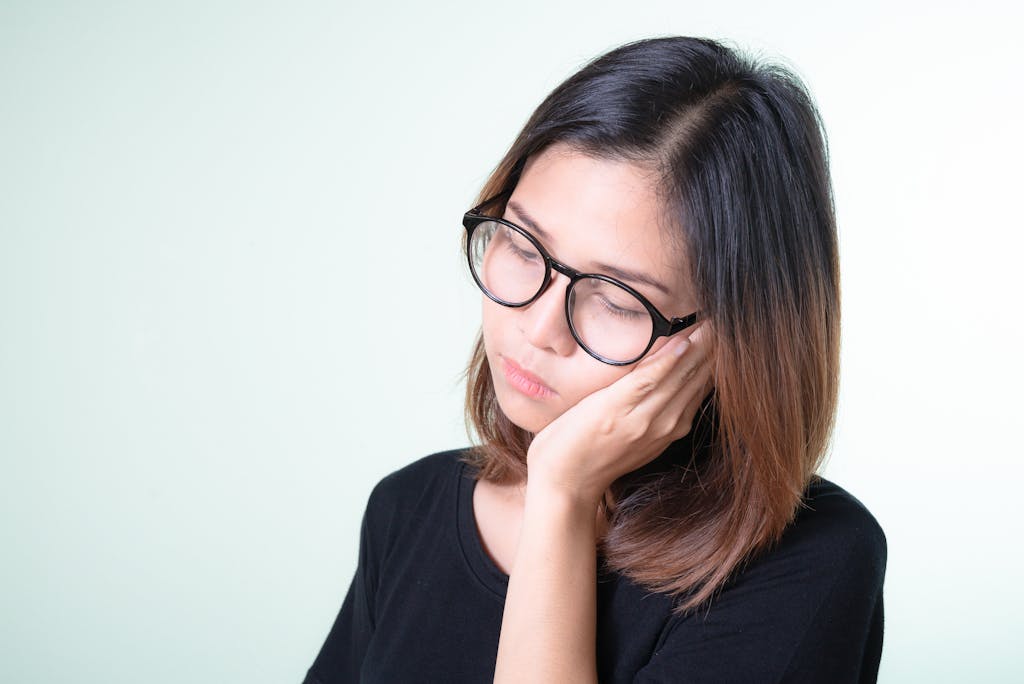 Portrait of a thoughtful young woman in glasses with hand on cheek against a neutral background.