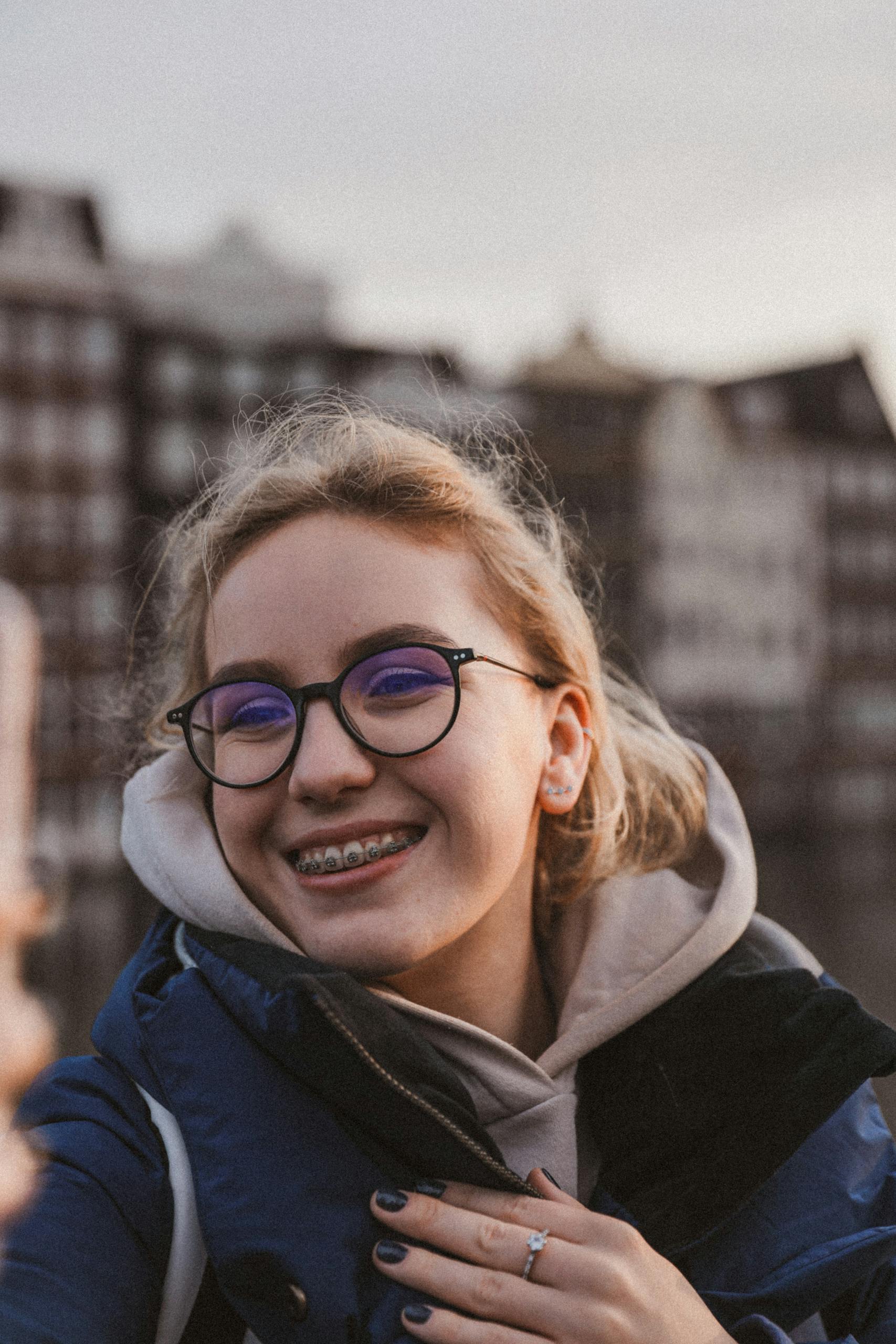 Smiling teenage girl with braces and glasses enjoying a moment outdoors, wearing a jacket.