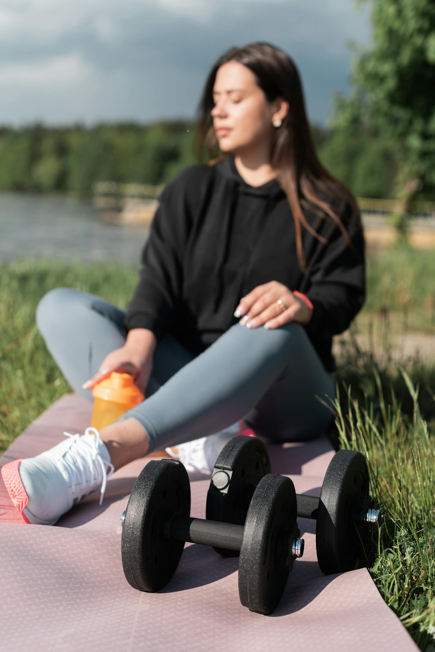 Young woman sits on a yoga mat with dumbbells outdoors, holding a water bottle, emphasizing wellness.